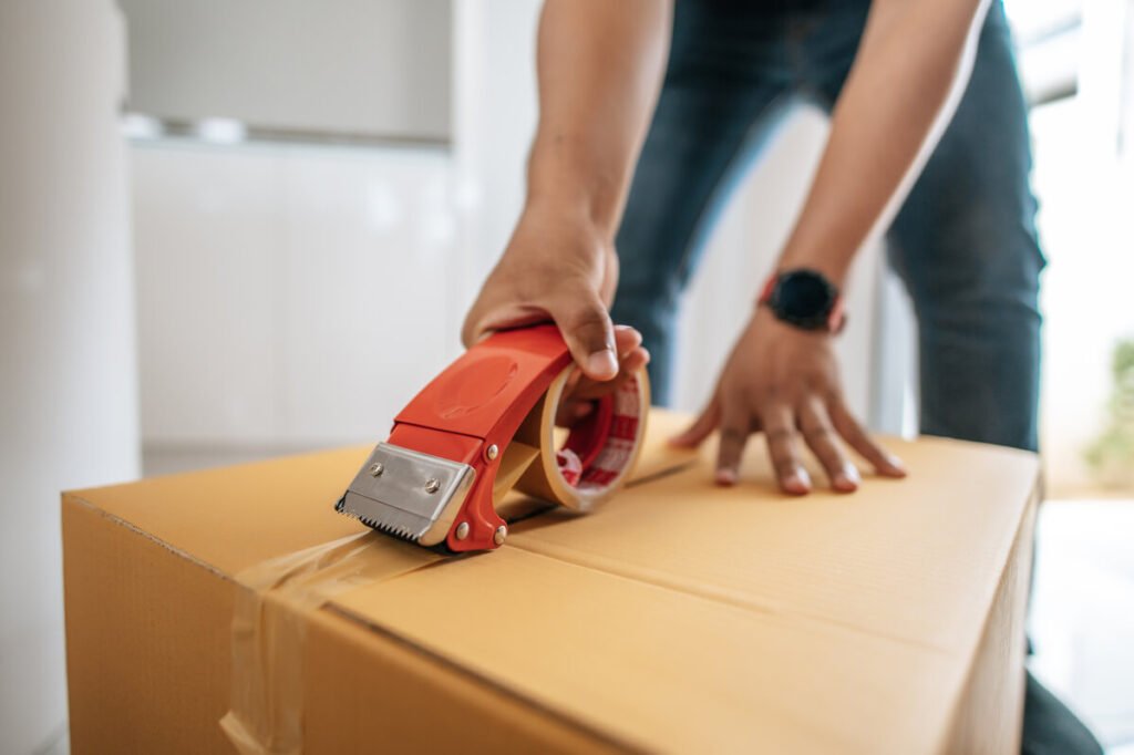 Déménagement : comment bien préparer son changement de logement ? 2 Personne fermant un carton de déménagement avec du ruban adhésif dans une pièce vide.